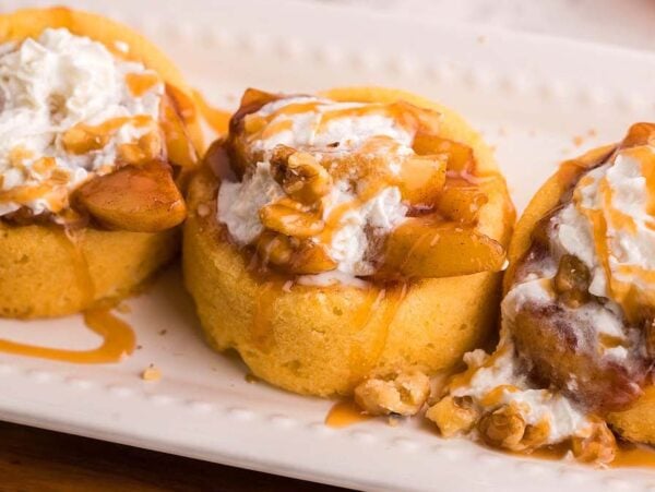 Three mini fireball apple shortcake dessert cups topped with whipped cream, caramel sauce, apple slices, and chopped walnuts make a delightful treat on a white plate.
