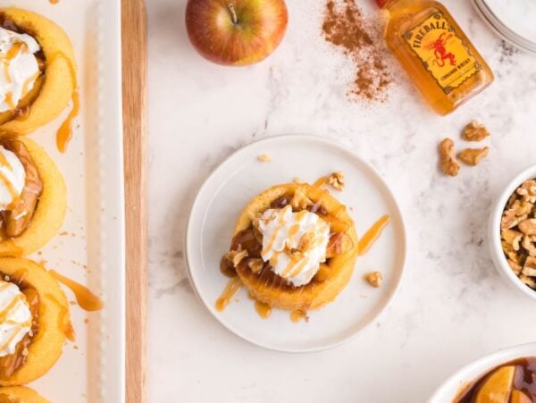 Mini sponge cake dessert cups topped with whipped cream, caramel, walnuts, and cinnamon apples soaked in Fireball, surrounded by more walnuts and a fresh apple.