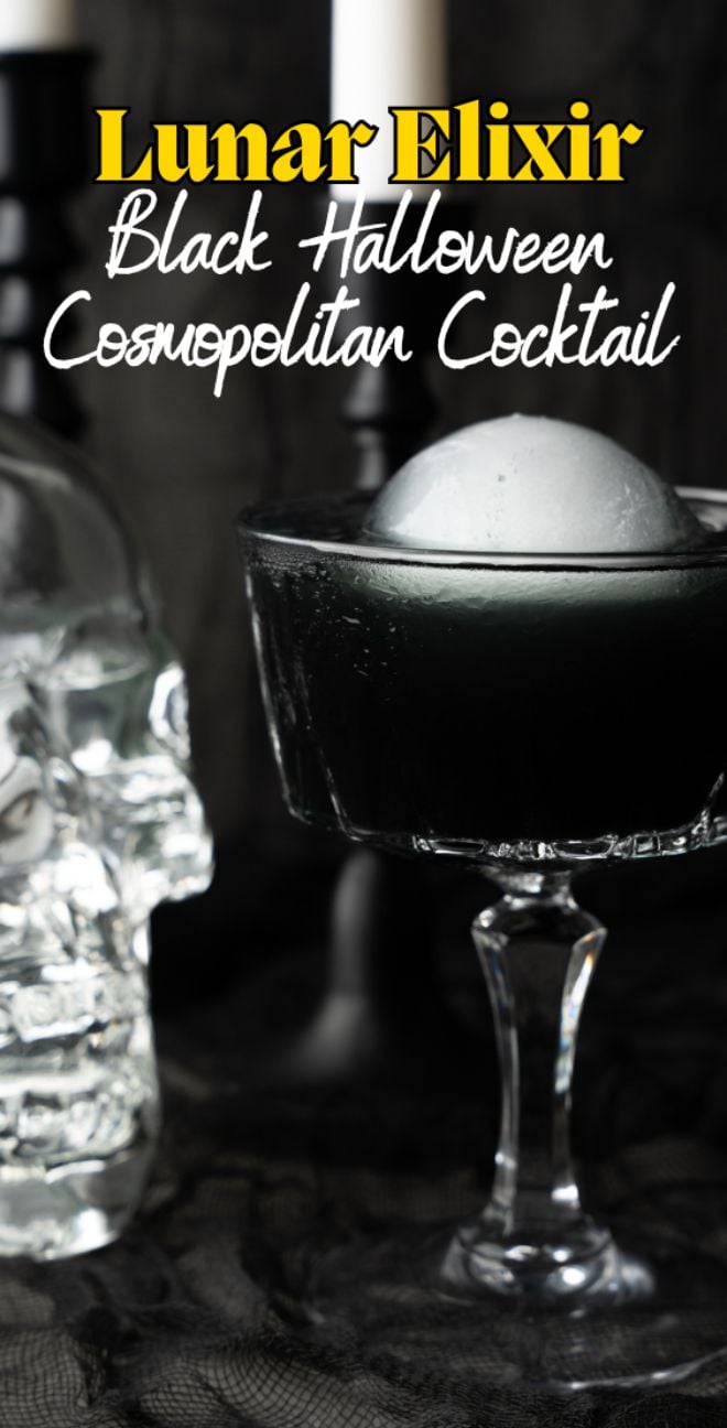 A Halloween themed drink: a black cocktail with an ice ball in a glass, set beside a glass skull and candles, surrounded by spooky Halloween decor.