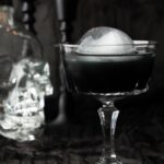 A black cocktail with an ice ball sits in a glass beside a skull-shaped bottle, creating the perfect Halloween vodka drink against a black background.