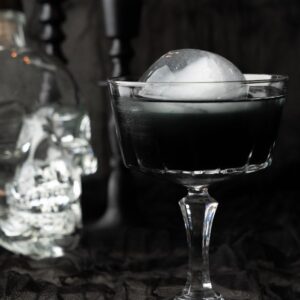 A black cocktail with an ice ball sits in a glass beside a skull-shaped bottle, creating the perfect Halloween vodka drink against a black background.