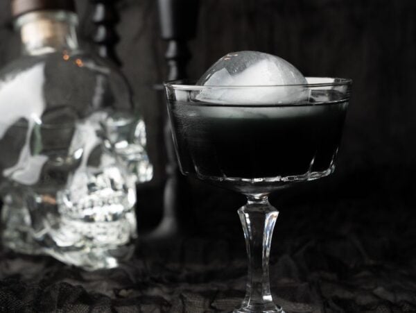 A black cocktail with an ice ball sits in a glass beside a skull-shaped bottle, creating the perfect Halloween vodka drink against a black background.