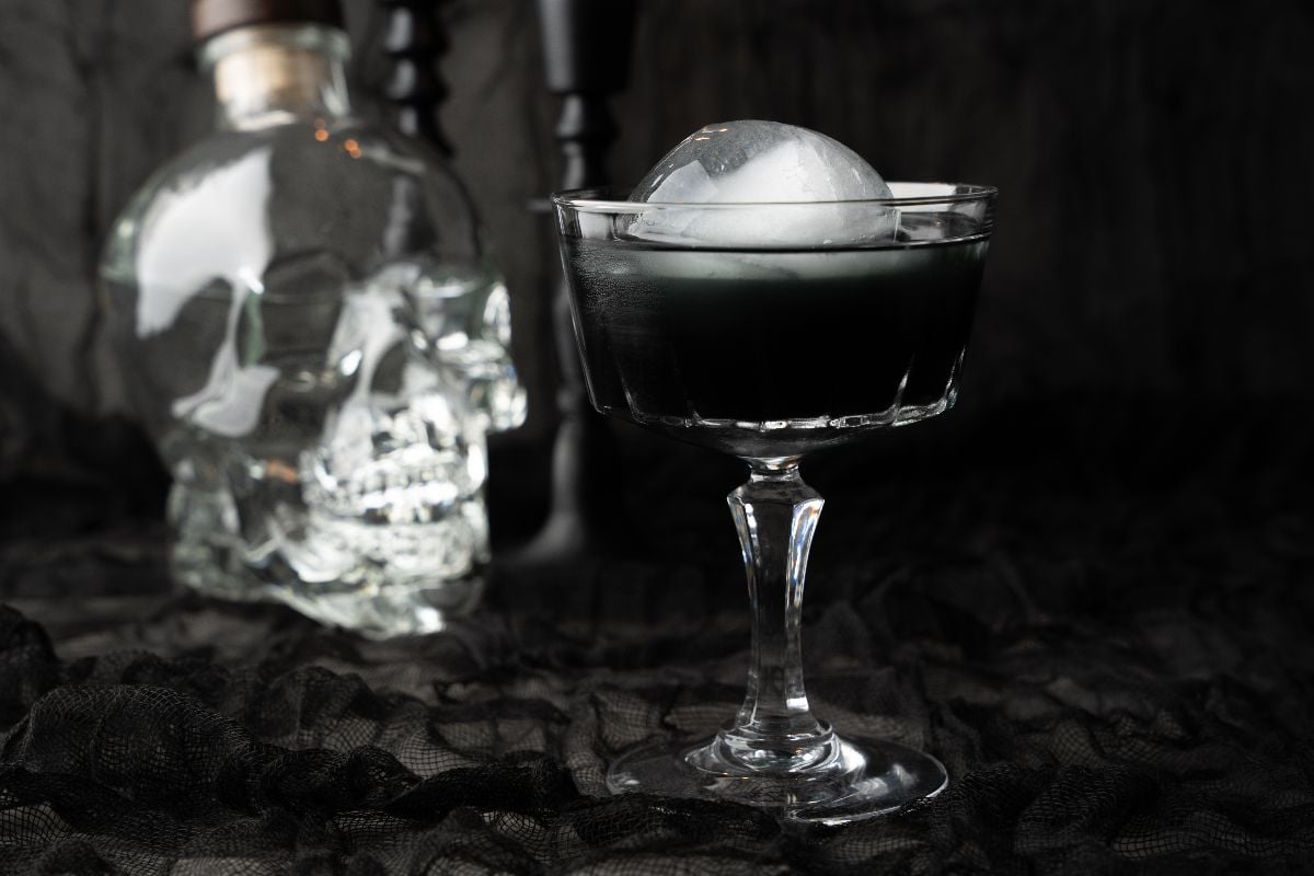 A Black Halloween Cosmopolitan Cocktail with a round ice sphere sits in a glass, beside a clear glass skull bottle on black fabric.