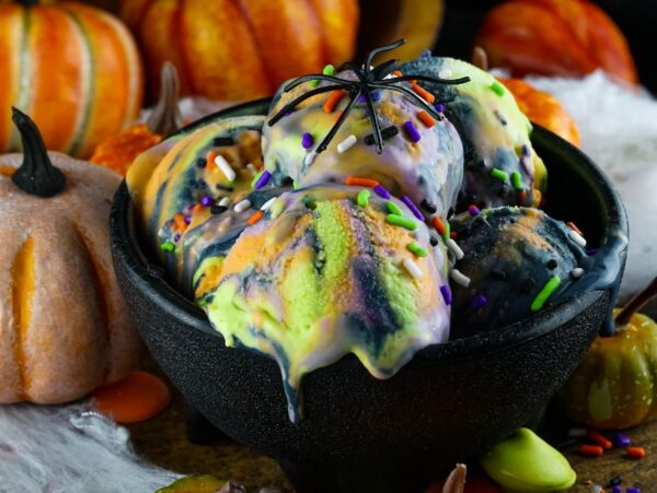 Black bowl of colorful No Churn Halloween Ice Cream with sprinkles, topped with a fake spider, surrounded by pumpkins—the perfect spooky ice cream for your frozen Halloween treats.