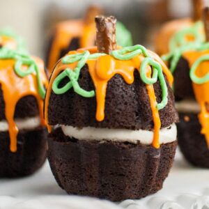 Chocolate cupcakes decorated like pumpkins with orange icing, green vines, and pretzel stick stems—perfect as a Thanksgiving or Halloween dessert or for learning how to make a pumpkin shaped cake.