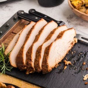 Sliced seasoned turkey breast on a black cutting board with a sprig of rosemary, showcasing the flavors of a boneless turkey breast in a slow cooker. Sides appear blurred in the background for an inviting meal scene.