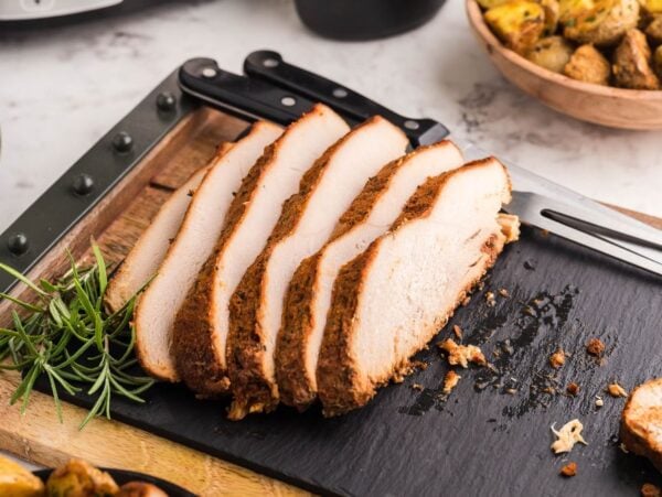 Sliced seasoned turkey breast on a black cutting board with a sprig of rosemary, showcasing the flavors of a boneless turkey breast in a slow cooker. Sides appear blurred in the background for an inviting meal scene.