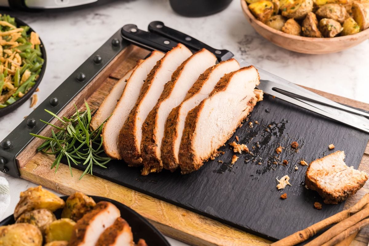 Sliced seasoned turkey breast on a black cutting board with a sprig of rosemary and kitchen knife nearby—perfect for trying an easy brine for turkey breast.