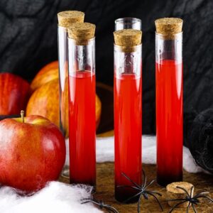 Four test tubes with red liquid and corks, apples, fake spiders, cobweb, and black cloth in a spooky setting—perfect for make ahead shots in a vial or syringe at your next Halloween party.
