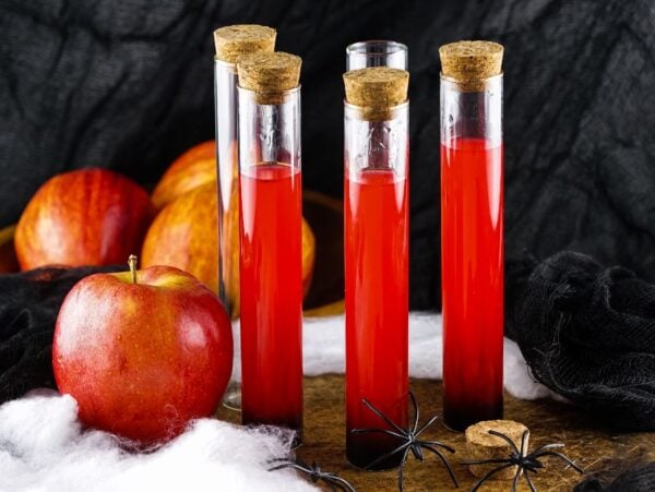 Four test tubes with red liquid and corks, apples, fake spiders, cobweb, and black cloth in a spooky setting—perfect for make ahead shots in a vial or syringe at your next Halloween party.