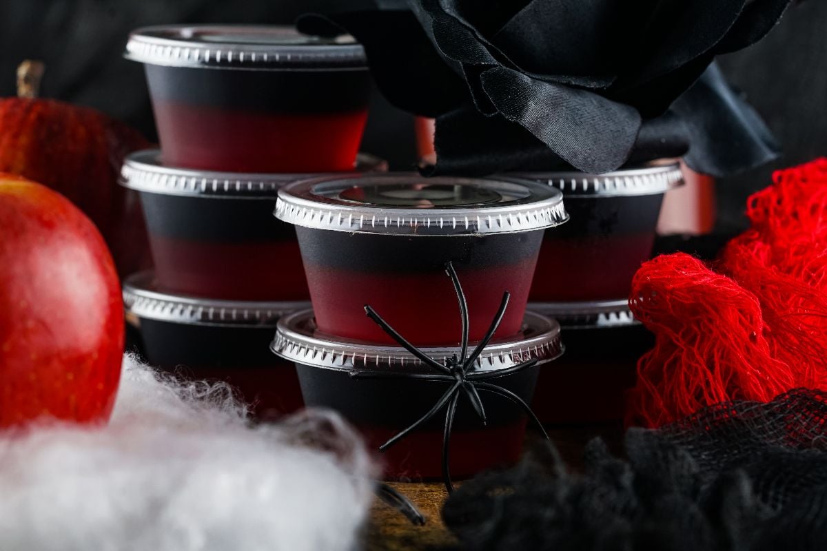 Stacked containers of red and black layered Vampire Kiss Jello Shots, with a fake spider and Halloween decorations nearby.