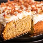 A slice of frosted Air Fryer Cornbread Cake with Honey Buttercream is lifted from the whole cake on a black plate, showcasing its delightful crumbled toppings and creamy Honey Buttercream frosting.