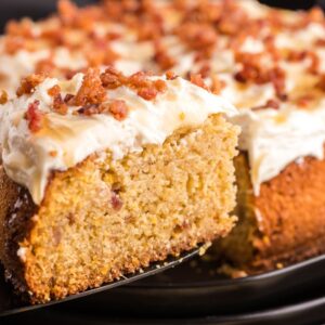 A slice of frosted Air Fryer Cornbread Cake with Honey Buttercream is lifted from the whole cake on a black plate, showcasing its delightful crumbled toppings and creamy Honey Buttercream frosting.