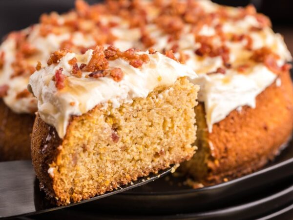 A slice of frosted Air Fryer Cornbread Cake with Honey Buttercream is lifted from the whole cake on a black plate, showcasing its delightful crumbled toppings and creamy Honey Buttercream frosting.
