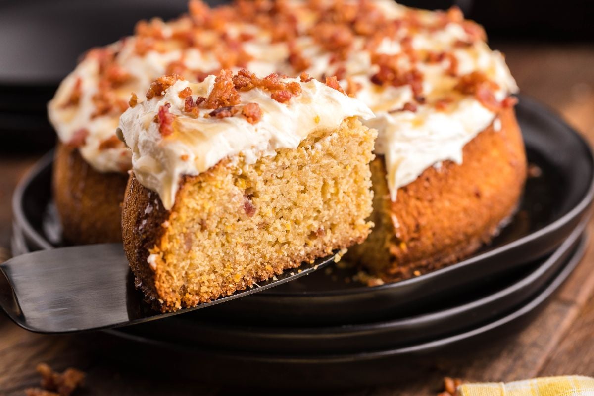 A slice of Air Fryer Cornbread Cake with Honey Buttercream frosting and crumbled topping is being served from a whole cake on a black plate, making for a unique cornbread dessert.