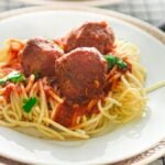 Spaghetti topped with gluten free air fryer meatballs, marinara sauce, and parsley on a white plate with a silver rim.
