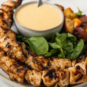 Cider grilled chicken skewers on a plate with fresh greens, roasted vegetables, and a bowl of creamy Gouda Cheese Sauce for dipping.