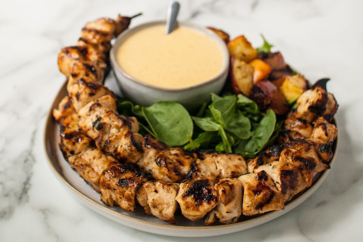 Apple Cider Chicken Skewers with Gouda Sauce served with greens, roasted vegetables, and a bowl of dipping sauce on a plate.
