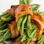 Bacon Wrapped Green Beans are stacked on a white plate, featuring oven roasted green beans wrapped in crispy bacon for a savory bite.