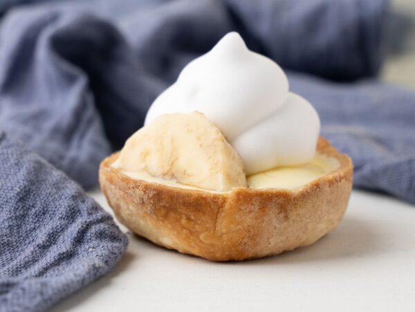 Mini Banana Cream Pie topped with whipped cream, made with pre-made pie crust and instant vanilla pudding, sits on a light surface with a blue cloth in the background.