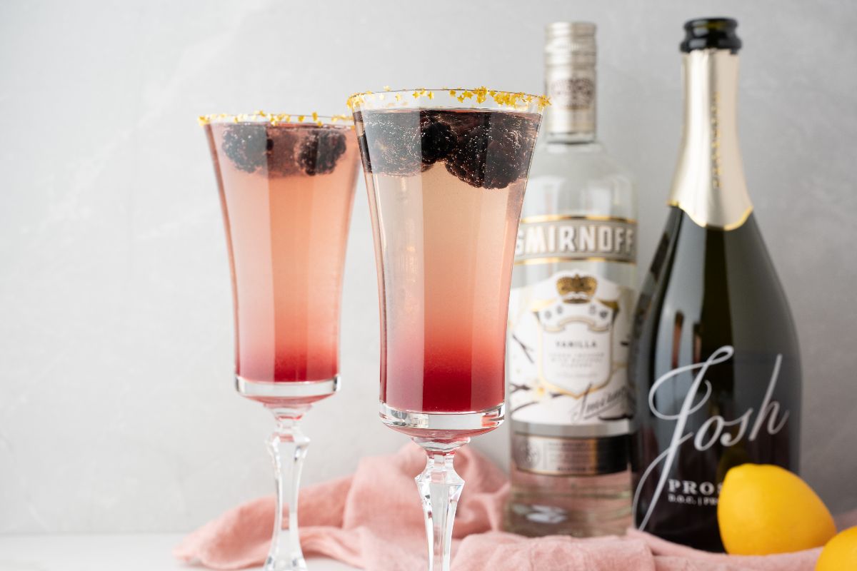 Two berry cocktails in flutes, made with Meyer lemon blackberry syrup, sit beside vodka and prosecco bottles, a lemon, and pink cloth—perfect as a festive Lemon Berry Prosecco Cocktail for your Christmas or New Year’s Eve celebration.