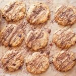 Twelve Peanut Butter Chocolate Chunk Cookies drizzled with chocolate on parchment, with bowls of melted chocolate, almonds, and a glass of milk.