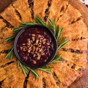 Round savory pastry arranged like a Christmas wreath with a bowl of cranberry dip in the center, garnished with rosemary sprigs—perfect for serving as a festive cranberry brie wreath appetizer.