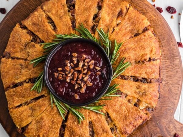 Round savory pastry arranged like a Christmas wreath with a bowl of cranberry dip in the center, garnished with rosemary sprigs—perfect for serving as a festive cranberry brie wreath appetizer.