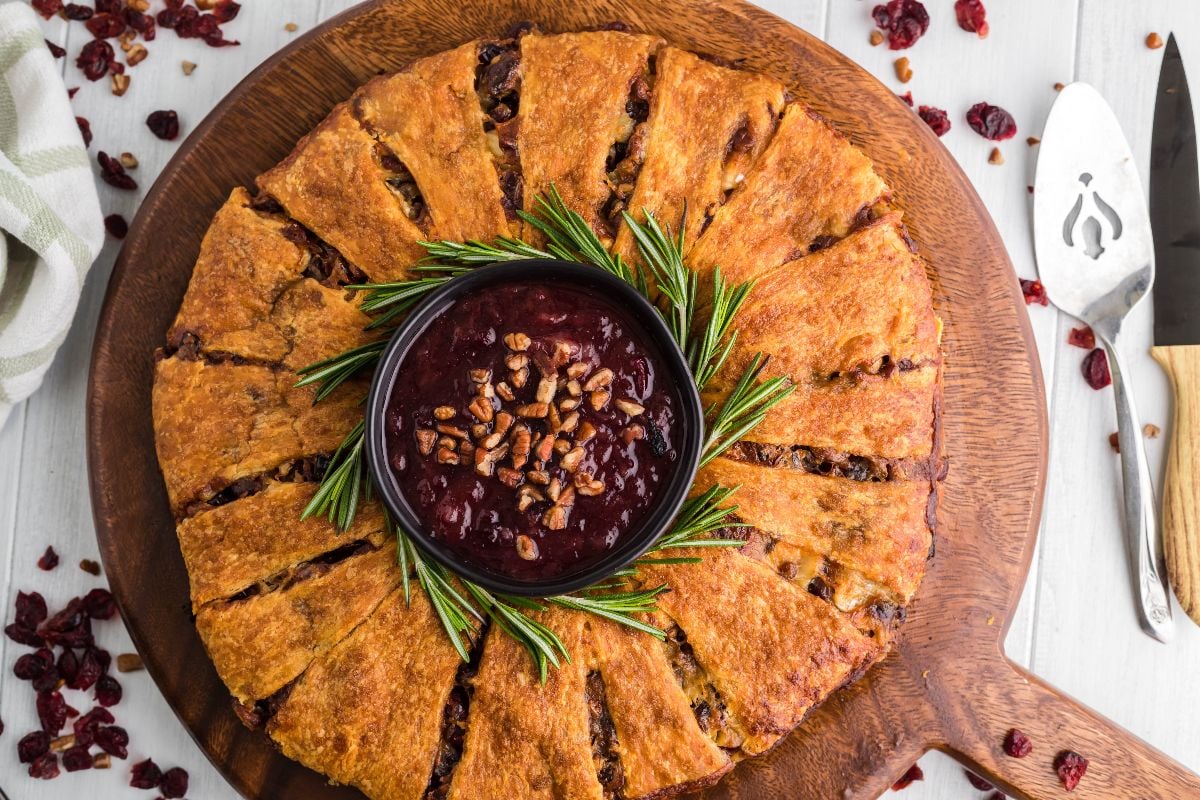 Round sliced Pineapple Cranberry Brie Wreath with a bowl of cranberry sauce and rosemary sprigs in the center, styled like a festive Christmas wreath on a wooden board.