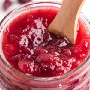 A glass jar filled with spiked cranberry sauce, with a wooden spoon inside, showcases this delicious cranberry sauce with bourbon and pineapple.