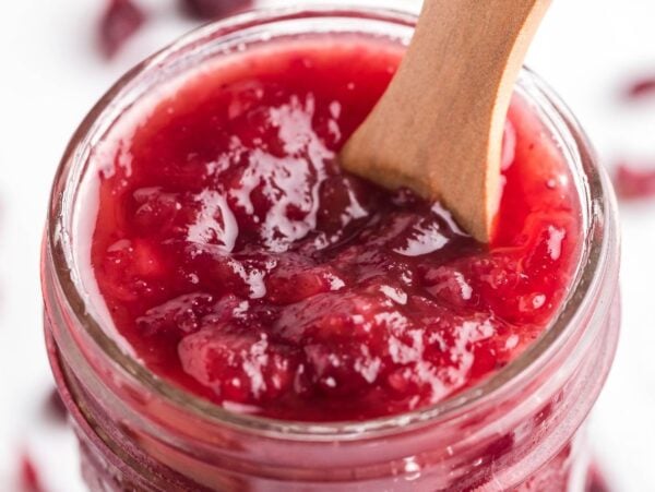 A glass jar filled with spiked cranberry sauce, with a wooden spoon inside, showcases this delicious cranberry sauce with bourbon and pineapple.