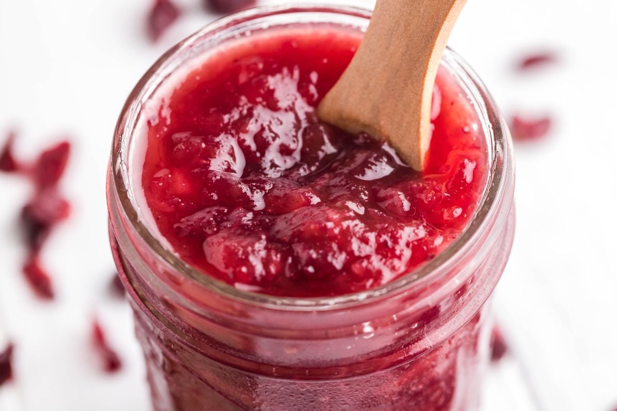 A glass jar of cranberry sauce with a wooden spoon inside it, on a white surface—perfect for serving cranberry bourbon relish at your next gathering.
