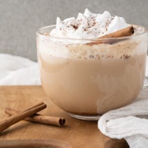 A glass mug of chai latte topped with whipped cream, cinnamon, and a cinnamon stick on a wooden board—a cozy DIY cinnamon dolce latte perfect for recreating café vibes at home.