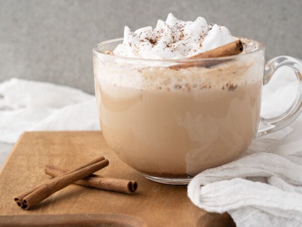 A glass mug of chai latte topped with whipped cream, cinnamon, and a cinnamon stick on a wooden board—a cozy DIY cinnamon dolce latte perfect for recreating café vibes at home.