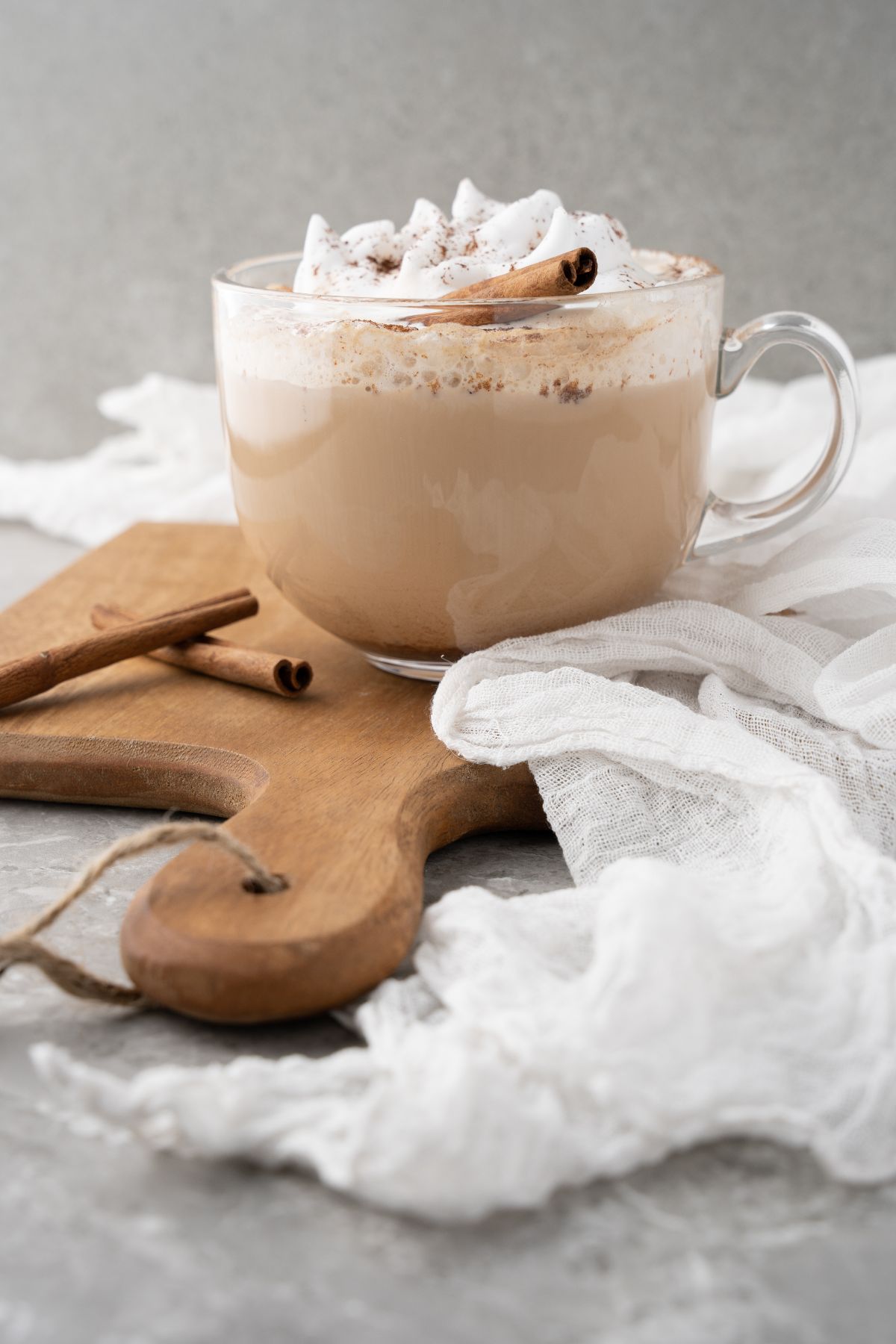 A glass mug of creamy latte topped with whipped cream and cinnamon, on a wooden board with a cinnamon stick—a true Starbucks Cinnamon Dolce Latte copycat experience.
