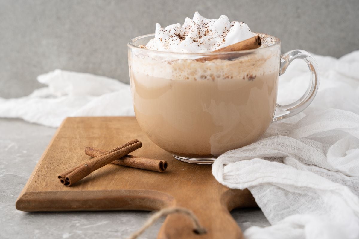 A glass mug of chai latte with whipped cream and cinnamon sticks on a wooden board, perfect for recreating a cozy DIY cinnamon dolce latte at home.