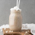 Iced coffee with whipped cream in a mason jar, two straws, on a wooden board with white chocolate chips—this is the perfect Starbucks Copycat Iced White Chocolate Mocha for your next DIY white chocolate mocha iced coffee treat.