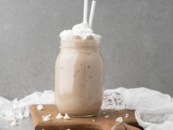 Iced coffee with whipped cream in a mason jar, two straws, on a wooden board with white chocolate chips—this is the perfect Starbucks Copycat Iced White Chocolate Mocha for your next DIY white chocolate mocha iced coffee treat.