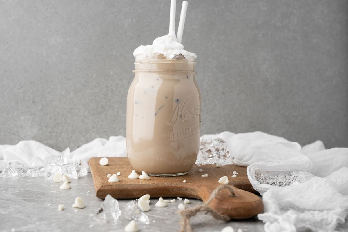 Homemade iced mocha with whipped cream and two straws in a mason jar on a wooden board, garnished with white chocolate chips—a perfect DIY white chocolate mocha iced coffee treat.