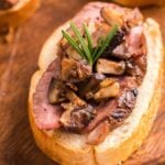 A slice of bread topped with roasted meat, mushrooms, and a sprig of rosemary on a wooden board—perfect for serving as Steak and Mushroom Bruschetta at your next gathering.