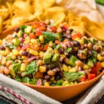 A bowl of colorful Texas Cowboy Caviar dip—also known as bean and corn salsa—sits in a basket, surrounded by crisp chips.