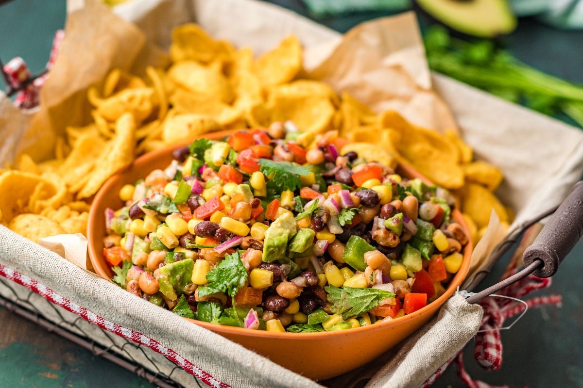 A bowl of colorful Texas Cowboy Caviar dip, loaded with beans and corn, served with tortilla chips on the side in a basket.