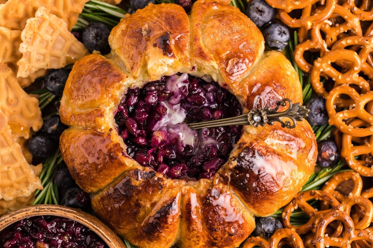 Round baked brie cheese dip with berry filling, surrounded by pretzels, waffle cookies, and fresh blueberries for a delightful blueberry brie appetizer.
