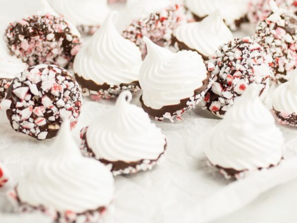 Dipped meringue kisses coated in chocolate and crushed peppermint, with candy canes on white parchment paper—a festive treat for christmas meringue cookie recipes.