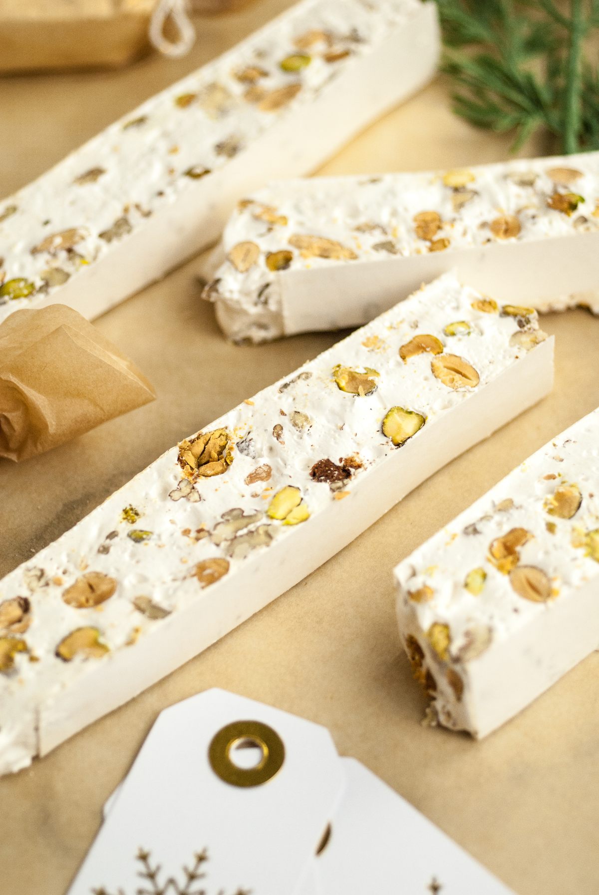 Rectangular slices of Italian Nut Nougat, a classic Italian nougat candy with mixed nuts, are arranged on parchment paper, surrounded by gift tags and festive greenery.