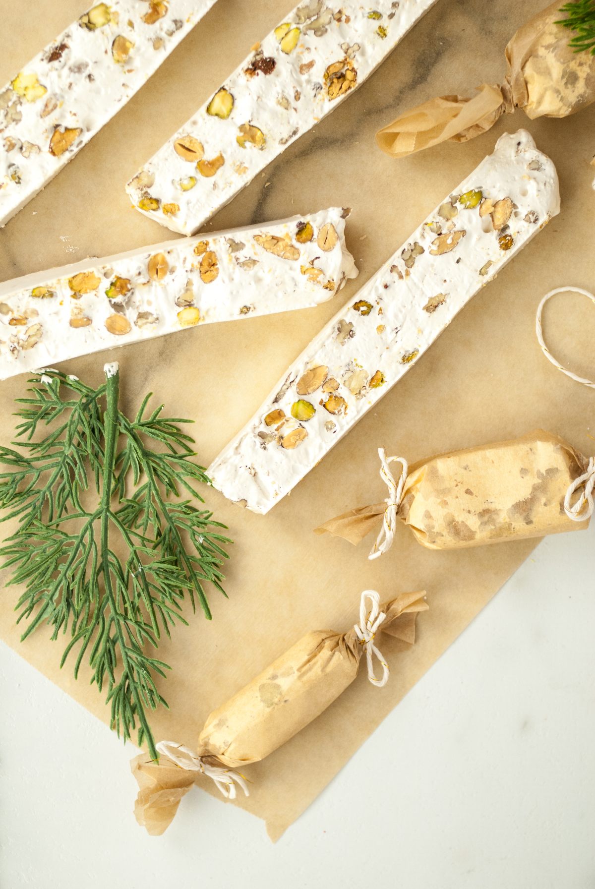 Italian nougat candy bars with roasted nuts and two wrapped treats rest on parchment paper, accented by green sprigs—a festive Christmas candy with roasted nuts.