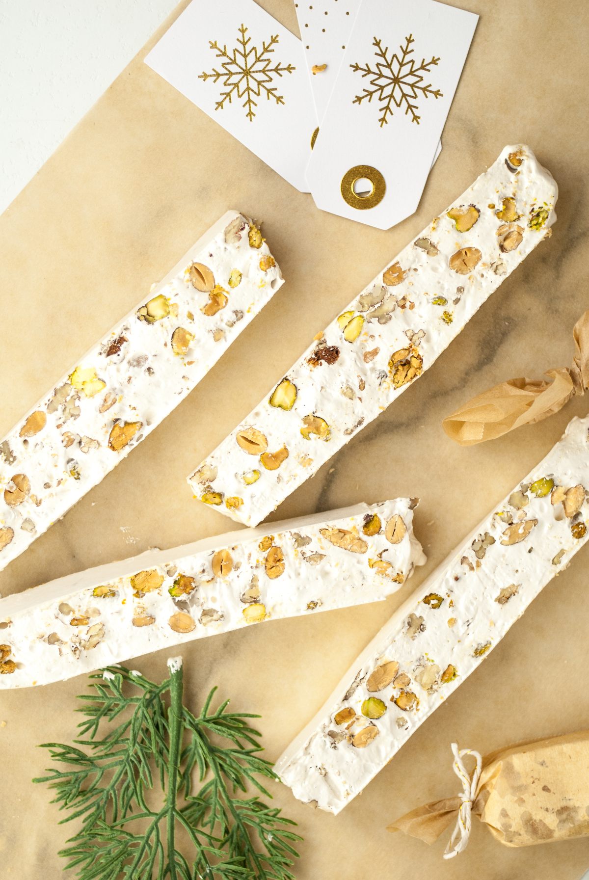 Four Italian Nut Nougat bars with nuts rest on parchment, surrounded by gift tags, rosemary, and wrapped italian nougat candy nearby.