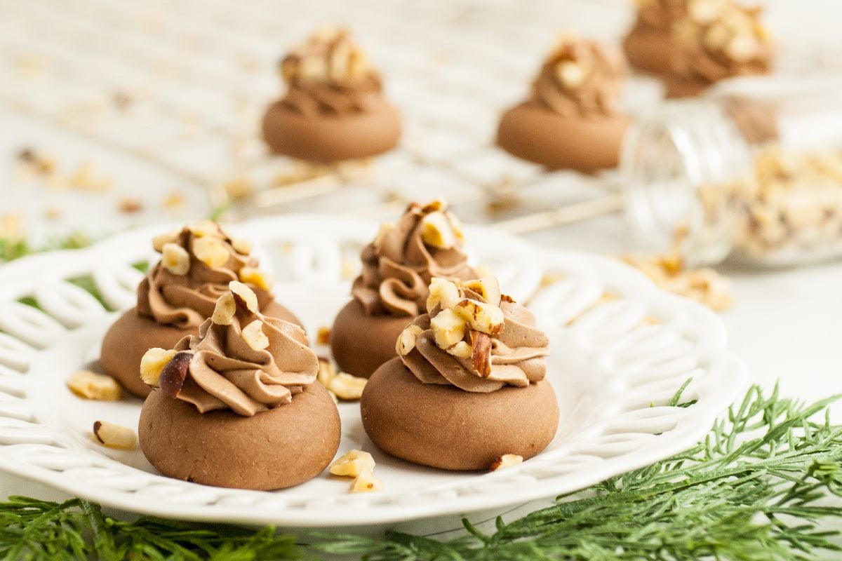 Chocolate cookies with swirls of frosting and chopped nuts on a white plate, inspired by Nutellotti—Italian Nutella cookies—with more cookies in the background. Perfect for those seeking recipes that use Nutella.