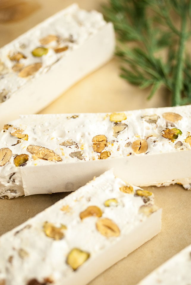 Close-up of nougat (torrone) bars with visible nuts on parchment paper and a green herb garnish in the background.