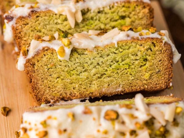 Close-up of sliced pistachio loaf cake with white icing and chopped nuts on a wooden board.